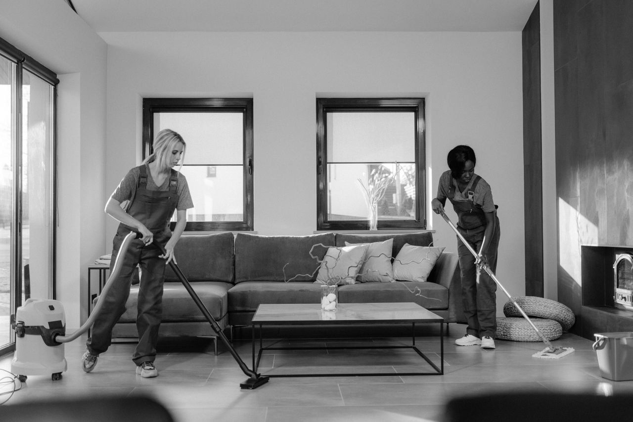 why-choose-us Two women in work clothes cleaning a modern living room using a vacuum and mop.